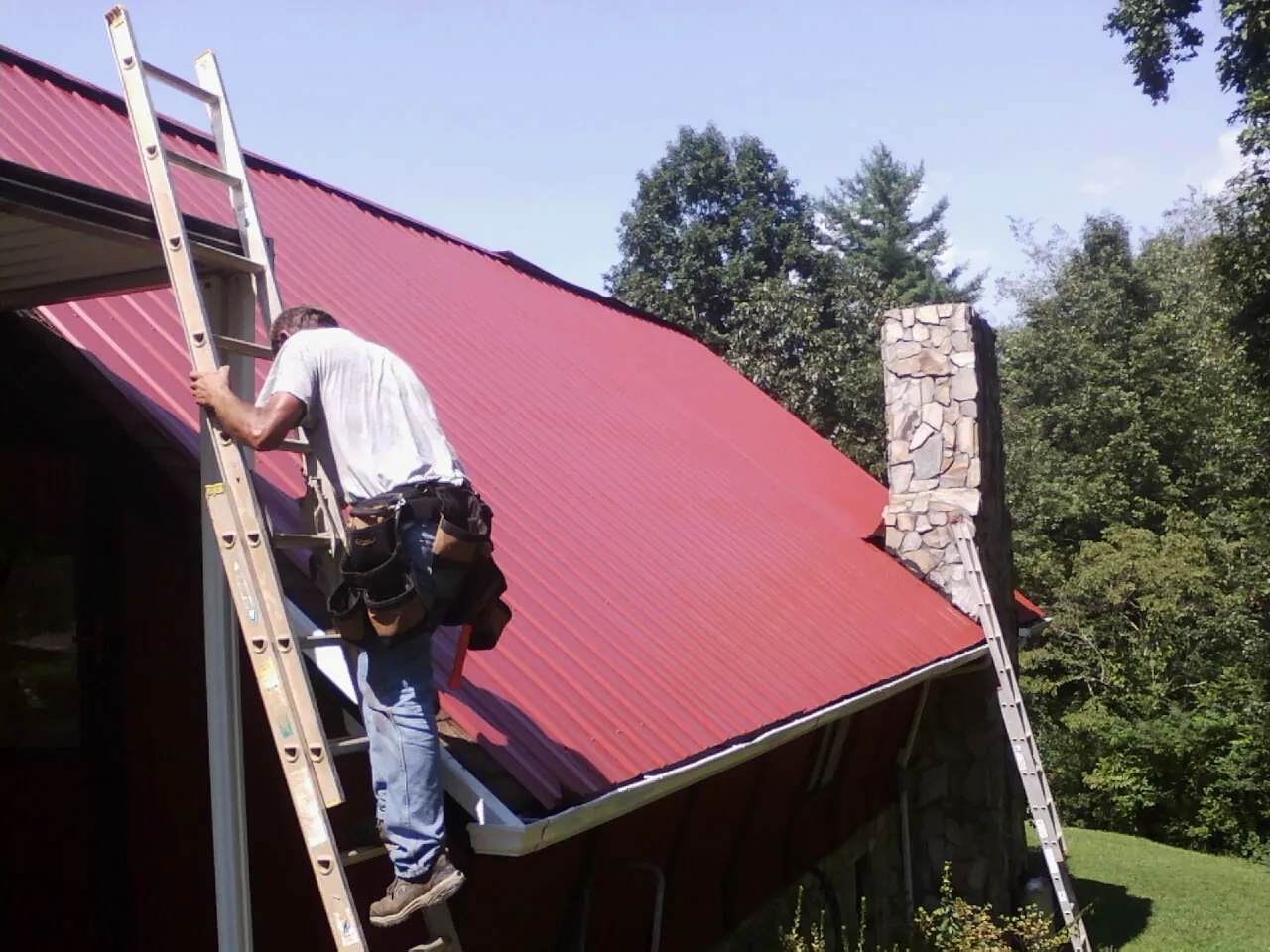 Roofer on a ladder beside a red metal roof and stone chimney — Soffit & Fascia Repair by Roofing Brothers in Renfrew